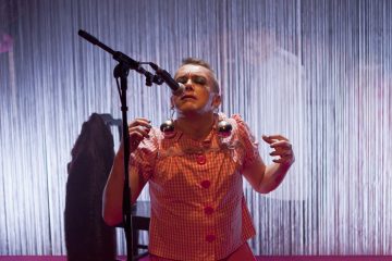 Vaaleanpunaiseen ruudulliseen mekkoon ja suuriin korvakoruihin pukeutunut esiintyjä seisoo mikrofonin edessä ja elehtii ilmeikkäästi. Taustalla on pystysuoria jousia tai verhoja ja lämmin näyttämövalaistus.