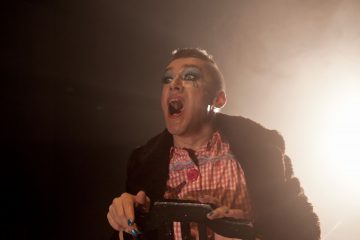 Henkilö, jolla on dramaattinen sininen silmämeikki, punainen huulipuna ja maalatut kynnet, pukeutuu ruutupaitaan ja tummaan turkistakkiin, pitelee tuolia ja laulaa tai huutaa ilmeikkäästi näyttämövalaistuksessa.