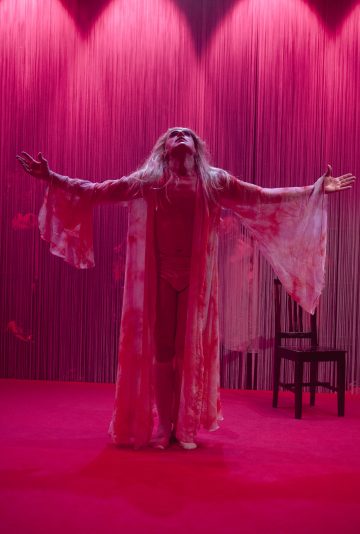 Pitkään, virtaavaan kaapuun pukeutunut henkilö seisoo näyttämön keskellä dramaattisessa vaaleanpunaisessa valaistuksessa, kädet ojennettuina ja pää taaksepäin kallistettuna, taustalla musta tuoli ja pystysuorat säikeet, jotka luovat kuvioidun taustan.
