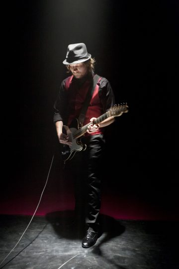 Mustaan hattuun, punaiseen liiviin ja mustiin vaatteisiin pukeutunut muusikko soittaa sähkökitaraa valonheittimen alla pimeällä lavalla.