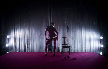 Muodonmukaiseen asuun pukeutunut henkilö seisoo punaisella lavalla, kamerasta poispäin, tuolin ja mikrofonitelineen vieressä, ja taustalla on dramaattinen taustavalo ja verhomaisia säikeitä.