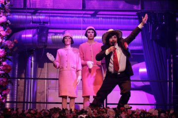 Pukuun ja olkihattuun pukeutunut mies elehtii tarmokkaasti lavalla kahden vaaleanpunaisiin pukuihin ja hattuihin pukeutuneen naisen edessä, ja heidän takanaan on dramaattinen violetti valaistus ja teollinen sisustus. Kukkia on näkyvissä näyttämön reunalla.