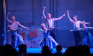 Kuusi tanssijaa vintage-tyylisissä puvuissa esittää dramaattisen, synkronoidun rutiinin lavalla siniviolettivärisessä valaistuksessa, taustalla EM-kirjaimet ja teollisuusympäristö.