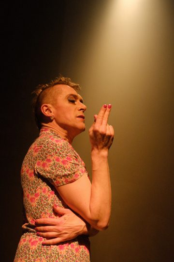 Kukkamekkoon ja meikkiin pukeutunut henkilö seisoo valonheittimen alla pitäen toista kättä rintakehänsä poikki ja toista kättä kasvojensa lähellä kaksi sormea kohotettuna, mikä herättää dramaattisen ja ilmeikkään poseerauksen.