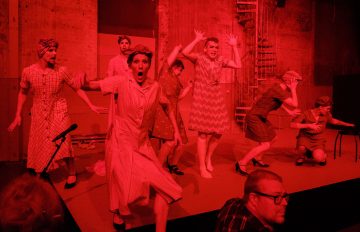 Ryhmä vintage-mekkoihin ja huiveihin pukeutuneita esiintyjiä esiintyy lavalla energisesti dramaattisen punaisen valaistuksen alla ja ilmaisee yllätystä ja jännitystä suurilla silmillä ja kohotetuin käsin. Etualalla näkyy yleisön jäsen.