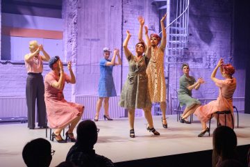 Kuusi vintage-tyylisiin mekkoihin pukeutunutta esiintyjää tanssii ja taputtaa lavalla violetin valaistuksen alla, ja jotkut istuvat tuoleilla ja toiset seisovat kesken liikkeen. Etualalla näkyy yleisön siluetteja.