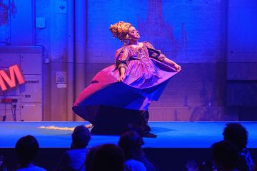 Esiintyjä, jolla on taidokas violetti ja kultainen puku ja päähine, poseeraa dramaattisesti lavalla sinisessä valaistuksessa, ja yleisö katsoo etualalta.