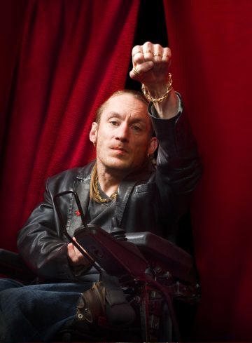 Pyörätuolissa istuva nahkatakkinen mies nostaa nyrkkiä punaisen verhon kehystämänä. Hänellä on lyhyet hiukset ja keskittynyt ilme, ja hänellä on kultaketju ja rannekoru.