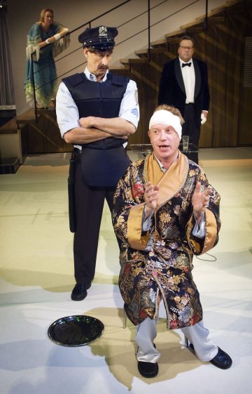 Kaapuun pukeutunut mies, jolla on sidottu pää, istuu puhumassa, kun hänen takanaan seisoo tiukka poliisi. Taustalla nainen seisoo portailla ja smokkiin pukeutunut mies katselee kohtausta.