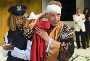 Kuvioituun kaapuun pukeutunut mies säätää päässään olevaa sidettä ja pitää kädessään punaista liinaa. Hänen vieressään poliisiksi pukeutunut nainen kirjoittaa muistikirjaan. Taustalla seisoo kaksi ihmistä.