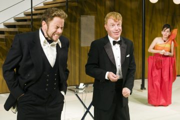 Lavalla esiintyy kolme juhlapukuista henkilöä. Etualalla seisoo kaksi smokkiin pukeutunutta miestä, joista toinen nyrpistelee silmät kiinni ja toinen hymyilee. Taustalla oranssiin pukuun pukeutunut nainen seisoo portaiden vieressä.