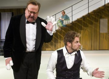 Lavalla esiintyy kaksi viralliseen asuun pukeutunutta miestä, joista toinen seisoo ja elehtii hansikkailla, kun taas toinen istuu ja katsoo muualle. Siniseen mekkoon pukeutunut nainen istuu taustalla portailla ja tarkkailee kohtausta.