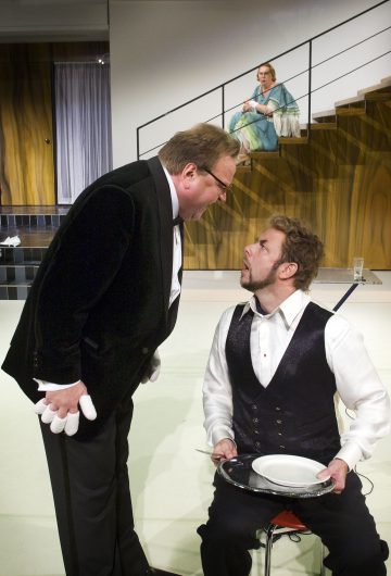 Mustaan pukuun pukeutunut mies nojaa aggressiivisesti kohti istuvaa miestä, jolla on lautanen ja tyhjä lautanen kädessään, kun taas sinipukuinen nainen istuu portailla taustalla ja tarkkailee kohtausta.