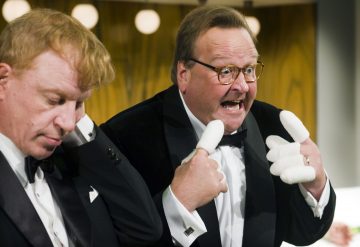 Kaksi smokkipukuista miestä sisätiloissa; toinen katsoo alaspäin käsi korvallaan, kun taas toinen osoittaa innoissaan itseään molemmilla peukaloillaan, jotka on sidottu valkoisella. Taustalla pyöreät valot ja puupaneeliseinä.