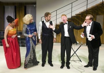 Viisi teatteripukuista henkilöä seisoo sisätiloissa. Yksi smokkipukuinen mies tekee liioiteltuja ilmeitä ja eleitä, ja muut katsovat uteliaina tai huolestuneina. Taustalla näkyy portaikko.