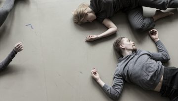 Kolme harmaisiin pukeutunutta ihmistä makaa vaalealla lattialla, kaksi vastakkain ja yksi osittain näkyvissä vasemmalla. Heidän asentonsa viittaa lepoon tai rentoutumiseen.