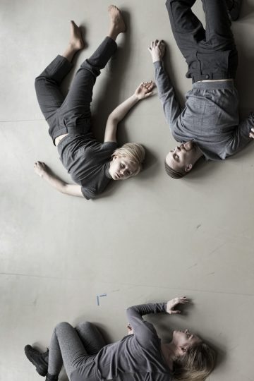 Kolme harmaisiin vaatteisiin pukeutunutta ihmistä makaa vaalealla lattialla muodostaen löysän kolmion. Kaksi makaa selällään, kun taas yksi on kyljellään. Heillä on ojennetut kädet, lähes toisiaan koskettavat kädet, ja he katsovat eri suuntiin.