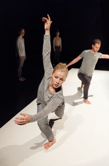 Harmaisiin vaatteisiin pukeutunut nainen esittää modernia tanssiliikkeitä toinen käsi kohotettuna, ja häntä ympäröi kolme muuta tanssijaa, joilla on samanlaiset asut valkoisella näyttämöllä tummalla taustalla.