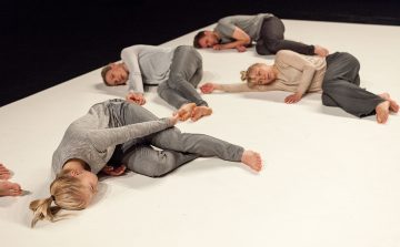 Neljä harmaisiin ja beigeihin lepotakkeihin pukeutunutta ihmistä makaa kyljellään valkoisella lattialla porrastetusti rivissä ja näyttää esittävän nykytanssia tai taiteellista liikettä.