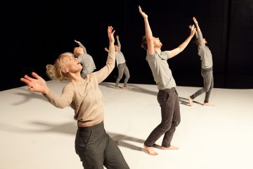 Viisi neutraalinvärisiin vaatteisiin pukeutunutta paljasjalkaista tanssijaa esiintyy valkoisella näyttämöllä, nostavat käsiään ja katsovat ylöspäin. Tausta on tumma, mikä kiinnittää huomion heidän ilmeikkäisiin liikkeisiinsä ja eleisiinsä.