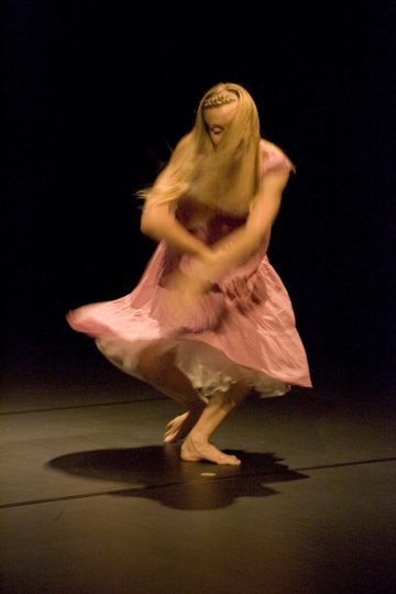 Vaaleanpunaiseen mekkoon pukeutunut henkilö tanssii energisesti tummalla lavalla, ja hänen pitkät vaaleat hiuksensa ja hameensa pyörivät liikkeestä.