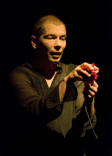 Hämärässä valaistuksessa seisoo henkilö, jolla on läpikuultava musta paita, ja joka katselee keskittyneesti molemmilla käsillään punaista kukkaa tummaa taustaa vasten.