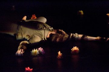 Henkilö makaa tummalla näyttämöllä toinen käsi ojennettuna, ja häntä ympäröivät hajallaan olevat värikkäät tekokukat. Pehmeä, dramaattinen valaistus korostaa ruumiinosia ja kukkia.