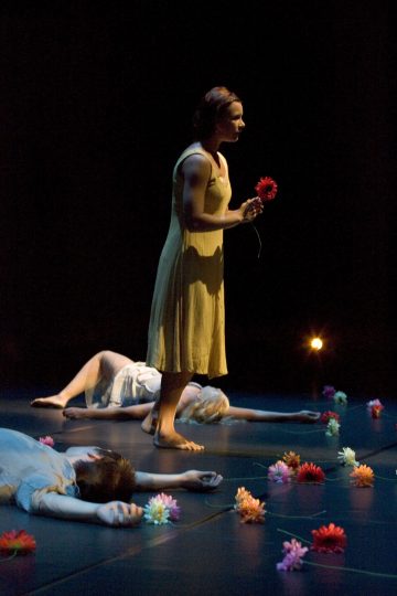 Keltaiseen mekkoon pukeutunut nainen seisoo paljain jaloin tummalla näyttämöllä, punainen kukka kädessään. Hänen ympärillään lattialla makaa kaksi ihmistä, joita ympäröivät hajallaan olevat värikkäät kukat. Valaistus on hämärä ja dramaattinen.