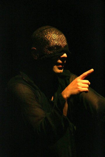 Tummiin, läpikuultaviin vaatteisiin ja silmät peittävään, kuvioituun naamariin pukeutunut henkilö osoittaa oikealla kädellään eteenpäin, joka on valaistu dramaattisesti tummaa taustaa vasten.