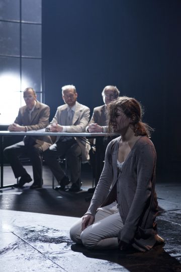 Nainen, jolla on märät hiukset ja vaatteet, polvistuu lattialle pöydän eteen, jossa istuu kolme pukumiestä, jotka tarkkailevat häntä tarkkaavaisesti. Kohtaus on hämärästi valaistu ja siinä on dramaattinen, synkkä tunnelma.