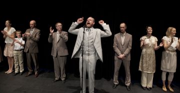 Ryhmä ihmisiä seisoo lavalla rivissä, useimmat taputtavat ja katsovat eteenpäin. Valkoiseen pukuun pukeutunut mies seisoo keskellä lavaa mikrofonin ääressä, nostaa nyrkkejään ja näyttää puhuvan tai laulavan kiihkeästi.