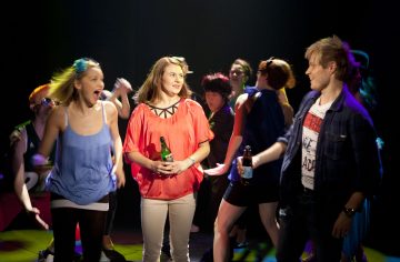 Ryhmä ihmisiä tanssii juhlissa. Etualalla punaisessa toppatakissa pulloa pitelevä nainen seisoo hymyilevän miehen ja nauravan naisen välissä, kun muut tanssivat tarmokkaasti taustalla värikkäiden valojen alla.