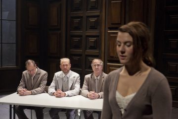 Etualalla seisoo nainen, joka katsoo alaspäin, kun taas kolme pukumiestä istuu pöydän takana ja tarkkailee häntä hämärässä huoneessa, jossa on tumma puupanelointi.