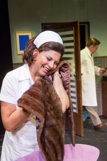 Sairaanhoitajaksi pukeutunut nainen hymyilee ja halajaa turkistakkia seisoessaan vaaleanpunaisen tuolin vieressä; valkoiseen takkiin pukeutunut mies on taustalla oven lähellä, kasvot poispäin.