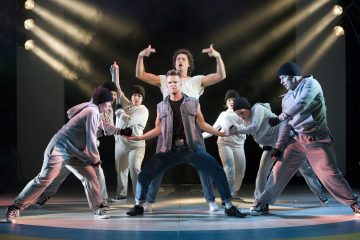 Ryhmä urheiluvaatteisiin pukeutuneita tanssijoita esiintyy lavalla dramaattisessa valaistuksessa. Yksi henkilö seisoo edessä polvet koukussa ja toinen takana kädet kohotettuna. Muut poseeraavat heidän ympärillään dynaamisissa asennoissa.