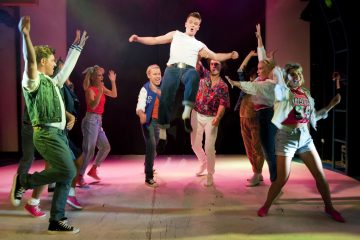 Ryhmä värikkäisiin 1980-luvun tyyliin pukeutuneita nuoria tanssii energisesti lavalla, ja yksi valkoiseen paitaan ja farkkuihin pukeutunut henkilö hyppää korkealle ilmaan keskellä kirkkaiden lavavalojen alla.