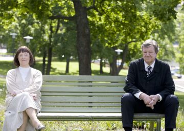 Vanhempi nainen ja mies istuvat erillään puiston vihreällä penkillä, molemmat katsovat neutraalilla ilmeellä eteenpäin. Taustalla on vihreitä puita ja ruohoa, jotka viittaavat rauhalliseen, aurinkoiseen päivään.