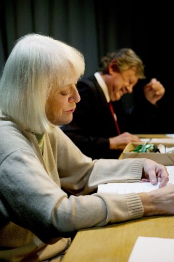 Vanhempi valkotukkainen nainen lukee pöydän ääressä asiakirjaa, ja hänen vieressään istuva pukuun pukeutunut vanhempi mies nauraa pää kääntyneenä ja käsi koholla. Molemmat ovat sisätiloissa lämpimässä valaistuksessa.