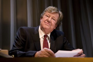 Tummaan pukuun ja punaiseen solmioon pukeutunut vanhempi mies istuu kirjoituspöydän ääressä papereita kädessään ja katsoo hieman ylöspäin mietteliäänä. Hänen takanaan on tumma verhotausta.