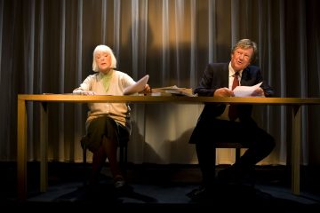 Vanhempi nainen ja mies istuvat puupöydän ääressä näyttämövalaistuksessa ja lukevat lehtiä. Taustalla on verho ja pöydällä on useita kirjoja. Molemmat näyttävät esiintyvän näytelmässä tai dramaattisessa lukemisessa.