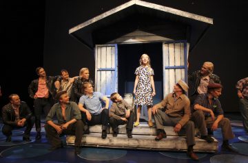 Kukkamekkoon pukeutunut nainen seisoo laulamassa sinivalkoisella puisella kuistilla, jota ympäröivät miehet, jotka istuvat ja katselevat häntä lavalla teatteriesityksen aikana.