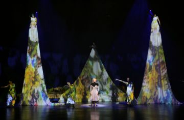 Näyttämöesitys, jossa keskeinen naispuolinen esiintyjä laulaa ja jota ympäröivät tanssijat ja kaksi korkeaa, kartionmuotoista rakennetta, jotka on valaistu värikkäillä, abstrakteilla valoprojektioilla dramaattisessa valaistuksessa.