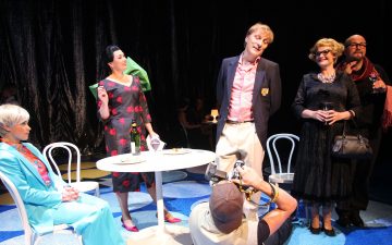 Viisi värikkääseen, tyylikkääseen asuun pukeutunutta ihmistä seisoo tai istuu näyttämöllä pienen valkoisen pöydän ympärillä. Yksi henkilö polvistuu edessä ja ottaa valokuvan, kun taas muut poseeraavat tai ovat vuorovaikutuksessa dramaattista, mustaa verhotaustaa vasten.