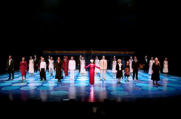 Suuri joukko ihmisiä seisoo etäällä toisistaan kirkkaasti valaistulla lavalla, jossa on sinisiä ympyräkuvioita. Keskellä seisoo henkilö, jolla on pitkä punainen mekko, kädet ojennettuina. Tausta on tumma.