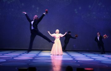 Vaaleanpunaiseen pukuun pukeutunut nainen seisoo näyttämön keskellä kädet ojennettuina, ja hänen ympärillään tanssii kolme smokkiin pukeutunutta miestä, joista yksi hyppii tarmokkaasti etualalla, ja kaikilla on kukkakimppu kädessään. Näyttämö on valaistu sinisen ja violetin sävyin.