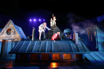 Värikkäisiin pukuihin pukeutuneet näyttelijät esiintyvät dramaattisesti hämärästi valaistussa, kattojen muotoon suunnitellussa lavastuksessa. Kaksi on etualalla, toinen kumartuneena liikkeessä, kun taas kaksi muuta seisoo taustalla, toinen nostelee kättä rekvisiitan avulla.