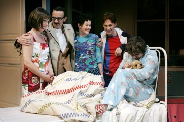 Viisi ihmistä kokoontui sängyllä istuvan henkilön ympärille, jolla oli pieni koira kädessään. Joukko vaikuttaa iloiselta, jotkut hymyilevät ja syleilevät. Kohtaus on sisätiloissa, ja värikkäät vaatteet ja vuodevaatteet luovat eloisaa tunnelmaa.