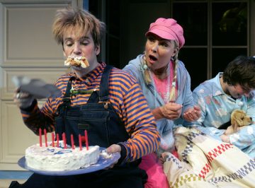 Haalareihin ja raidalliseen paitaan pukeutunut henkilö syö kakkua sotkuisesti, kuorrutus kasvoillaan ja pitää kädessään kynttiläkakkua. Toinen vaaleanpunaisessa lippiksessä oleva henkilö näyttää yllättyneeltä, ja kolmas henkilö istuu pehmoeläimen ja huovan kanssa.