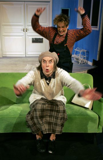 Vanhempi nainen, jolla on ruudullinen hame ja beige pusero, istuu vihreällä sohvalla kädet ojennettuina ja yllättynyt ilme, kun nuorempi raitapukuinen henkilö hyppii energisesti hänen takanaan. Valkoinen käsilaukku istuu sohvalla.