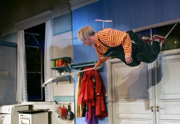 Punaraidalliseen paitaan ja haalariin pukeutunut henkilö leijuu vaakatasossa ilmassa propellihattu päässään, luuta kädessään värikkäässä keittiössä, lähellä valkoisia ovia ja ripustettuja takkeja.